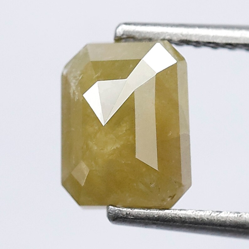May include: A yellow-green emerald cut diamond with a faceted surface. The diamond is held in place by a pair of tweezers.