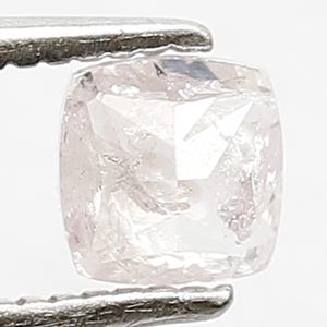 May include: A pale pink cushion-cut gemstone with a faceted surface, held by silver-toned metal tweezers. The stone displays a translucent quality with internal light reflections and sharp edges, showcasing a square shape with rounded corners.