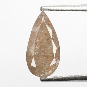 Puede incluir: Un diamante suelto en forma de pera con un tono marrón claro, color champán, y con inclusiones internas visibles. La piedra preciosa presenta una superficie facetada que refleja la luz, sujeta por unas pinzas metálicas de color plateado.