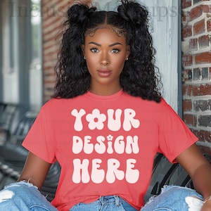 May include: A coral-colored t-shirt with the text "YOUR DESIGN HERE" in a white, retro font. The shirt has short sleeves and a crew neck. The model is wearing blue jeans.