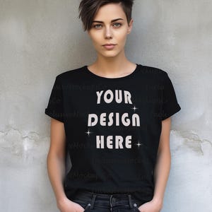 May include: Black t-shirt with the text "YOUR DESIGN HERE" in white, with star accents. The shirt has rolled-up sleeves and is paired with black jeans. The model is standing in front of a gray wall.
