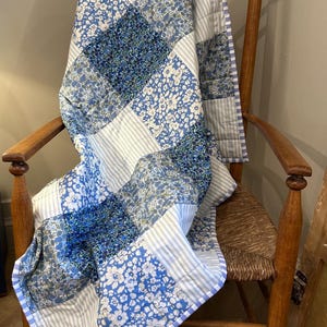 May include: A patchwork quilt featuring alternating squares of blue and white floral patterns and light blue and white striped fabric. The quilt is finished with a blue and white chequered border and displays visible stitching throughout the design.