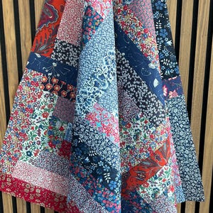 May include: A patchwork quilt featuring a variety of fabric patterns in shades of red, navy blue, light blue, and white. The design consists of rectangular and diagonal strips sewn together, showcasing floral, paisley, and geometric prints throughout.