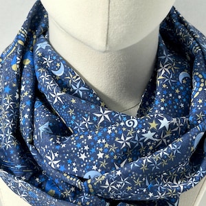 May include: A navy blue infinity scarf featuring a celestial pattern of white, light blue, and gold stars, moons, and other celestial elements. The scarf is draped around a mannequin head, highlighting its design and texture.