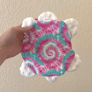 May include: A plush turtle-shaped baby washcloth with a tie-dye shell in pink, teal, and white. The turtle's head and limbs are made of white terry cloth. The washcloth is held against a neutral wall.