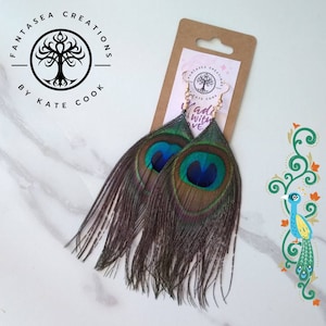 May include: A pair of long peacock feather earrings with gold hooks. The feathers are green, blue, and brown with a fringe of smaller feathers. The earrings are labeled "Fantasia Creations by Kate Cook" and "Made with Love".