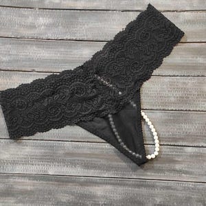 May include: Black lace thong underwear with a floral lace waistband. A string of white pearls is threaded through the thong section. The underwear is displayed on a grey wooden surface. The lace detailing is intricate.
