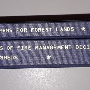 May include: Two blue book spines with white text. The top spine reads "Forest Lands" and the bottom spine reads "Fire Management Decision-Making".