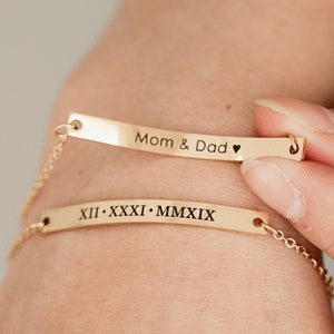 May include: Two gold chain bracelets with engraved bars. The top bar reads "Mom & Dad" with a heart symbol. The bottom bar reads "XII. XXXI. MMXIX".