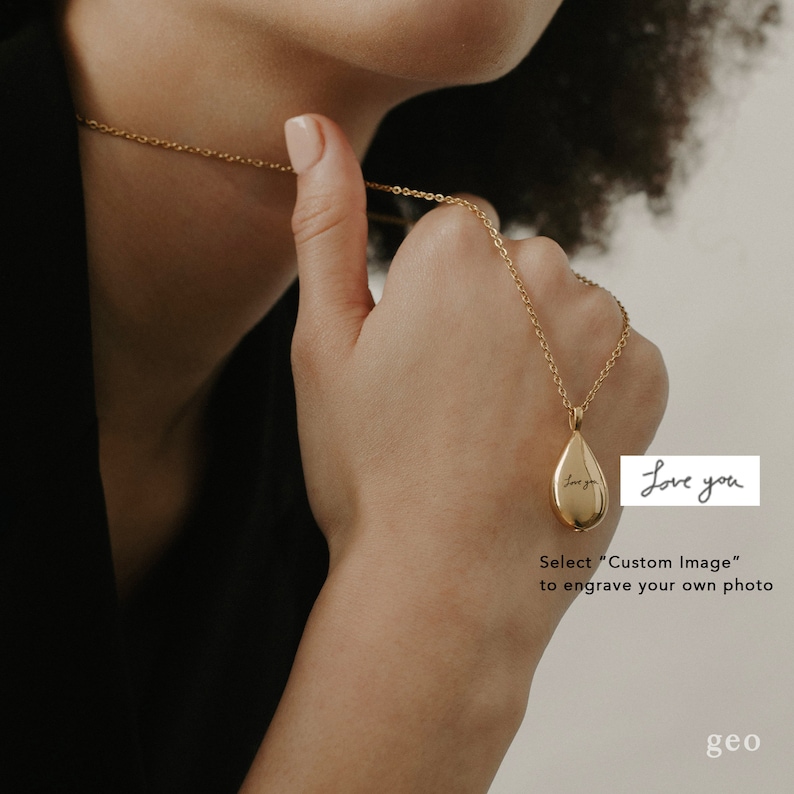 May include: A gold teardrop-shaped pendant necklace with a delicate chain. The pendant has the words "Love you" engraved on it. The necklace is being held by a person's hand.