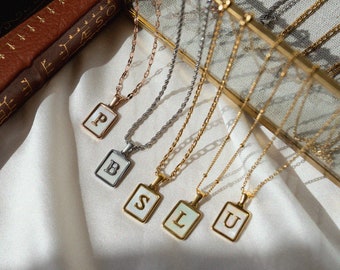 May include: Five gold necklaces with different initial charms, including P, B, S, L, and U. The necklaces are displayed on a white surface with a brown book in the background.