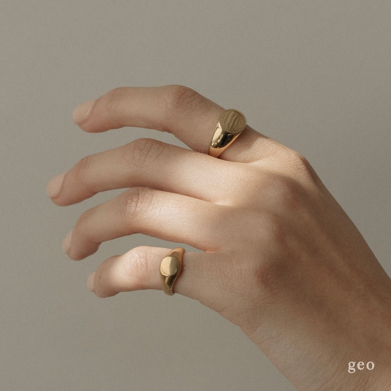Op de afbeelding: Twee gouden signet ringen op een hand. De ringen zijn ovaalvormig en hebben een gladde, gepolijste afwerking.