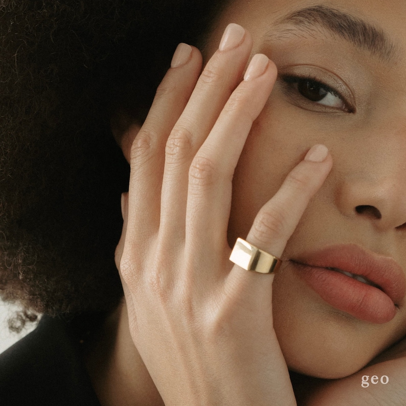 Puede incluir: Un anillo rectangular de oro usado en un dedo. El anillo es grueso y tiene una superficie lisa y pulida.