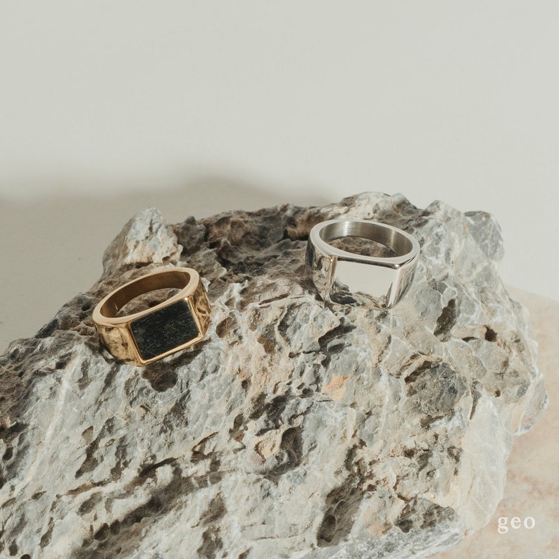 Puede incluir: Dos anillos sobre una roca gris. Un anillo es de oro con una piedra negra y el otro es de plata con una forma rectangular hueca.