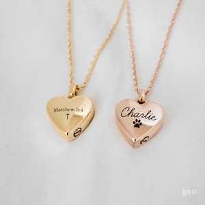May include: Two gold heart-shaped necklaces with engraved inscriptions. One necklace has the inscription "Matthew 5:4" and a cross. The other necklace has the inscription "Charlie" and a paw print.