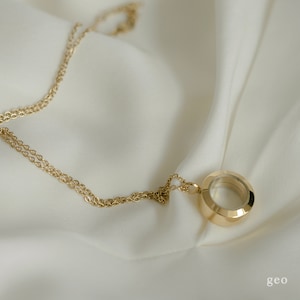 以下が含まれることがあります： 金色のネックレス。円形のペンダントが付いており、ファセットデザインと透明な中心部があります。繊細なチェーンは白くシルキーな生地の上にあります。「geo」の文字が右下にあります。