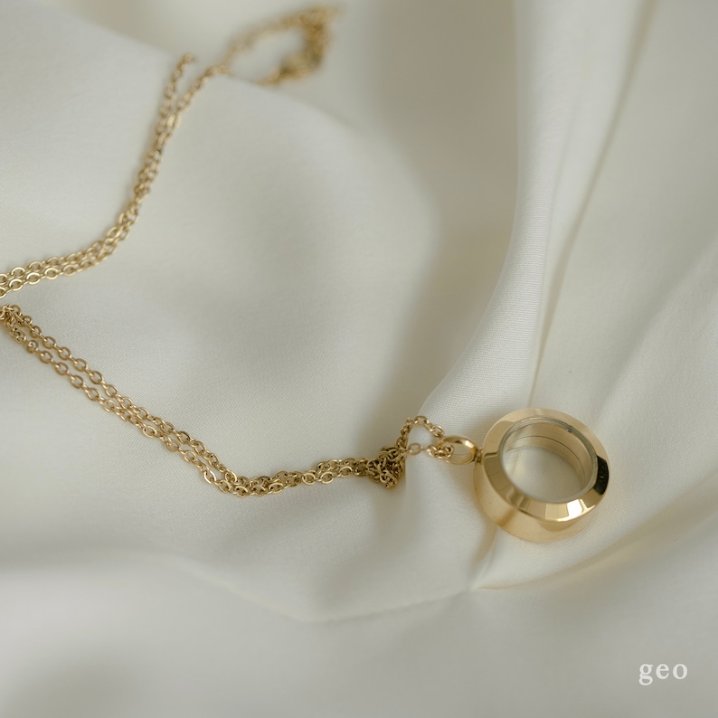 以下が含まれることがあります： 金色のネックレス。円形のペンダントが付いており、ファセットデザインと透明な中心部があります。繊細なチェーンは白くシルキーな生地の上にあります。「geo」の文字が右下にあります。