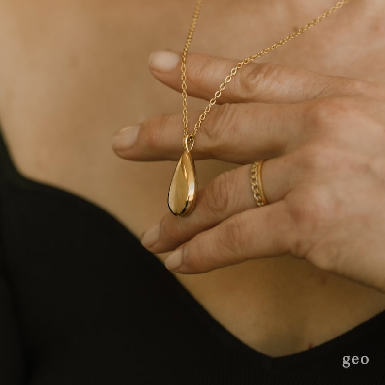 May include: A gold-coloured teardrop pendant necklace. The pendant is smooth and reflective, suspended from a delicate chain. A gold ring with a chain-like design is also visible. The necklace is held by a hand, with the word "geo" in the bottom right corner.
