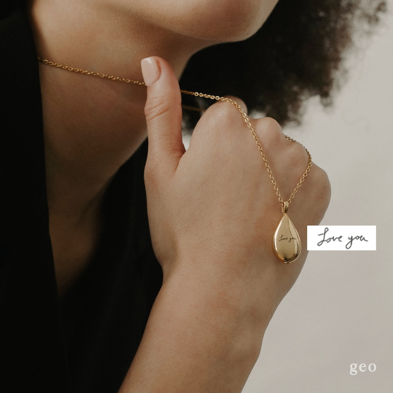 May include: A gold-coloured teardrop pendant necklace with the inscription "Love you". The necklace is held by a person wearing a black top. The pendant hangs from a delicate chain.