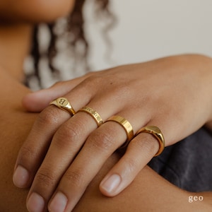 Op de afbeelding: Vier gouden ringen aan een hand. De ringen zijn allemaal in verschillende stijlen. Een ring heeft de letter "B" gegraveerd. Een andere ring heeft het Romeinse cijfer "XIX" gegraveerd. De andere twee ringen zijn gladde banden.