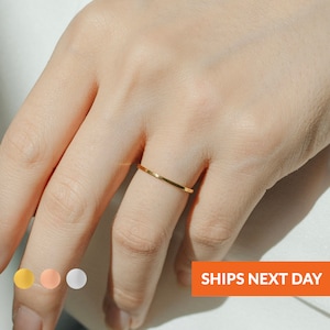 May include: A thin gold band ring on a finger. The ring is simple and delicate. The ring is available in gold, rose gold, and silver.