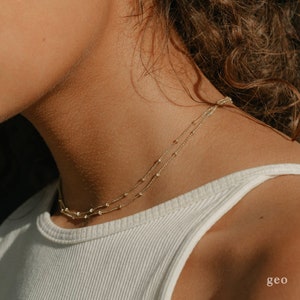 May include: A gold chain necklace with small beads, layered on a person's neck. The necklace is worn over a white tank top.