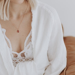 May include: A woman wearing a white robe with a delicate lace bralette underneath. She is wearing a gold necklace with a small red gemstone pendant.