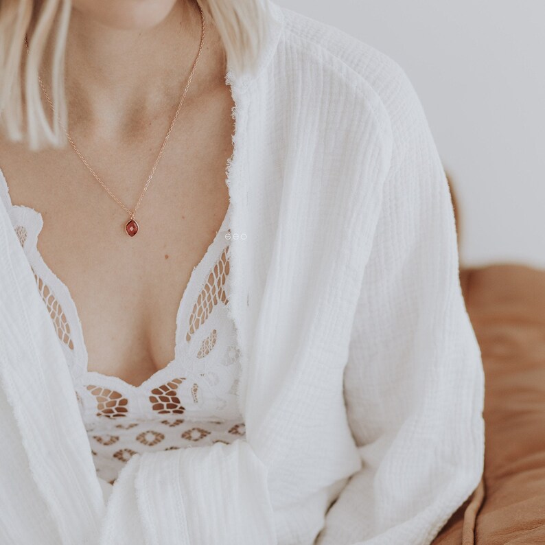 May include: A woman wearing a white robe with a delicate lace bralette underneath. She is wearing a gold necklace with a small red gemstone pendant.
