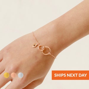 May include: A gold chain bracelet with two interlocking circles and a small leaf charm with a letter 'K' on it. The bracelet is shown on a person's wrist.