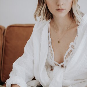 May include: A woman wearing a white shirt with a lace top underneath. She is wearing a gold necklace with a green stone pendant.