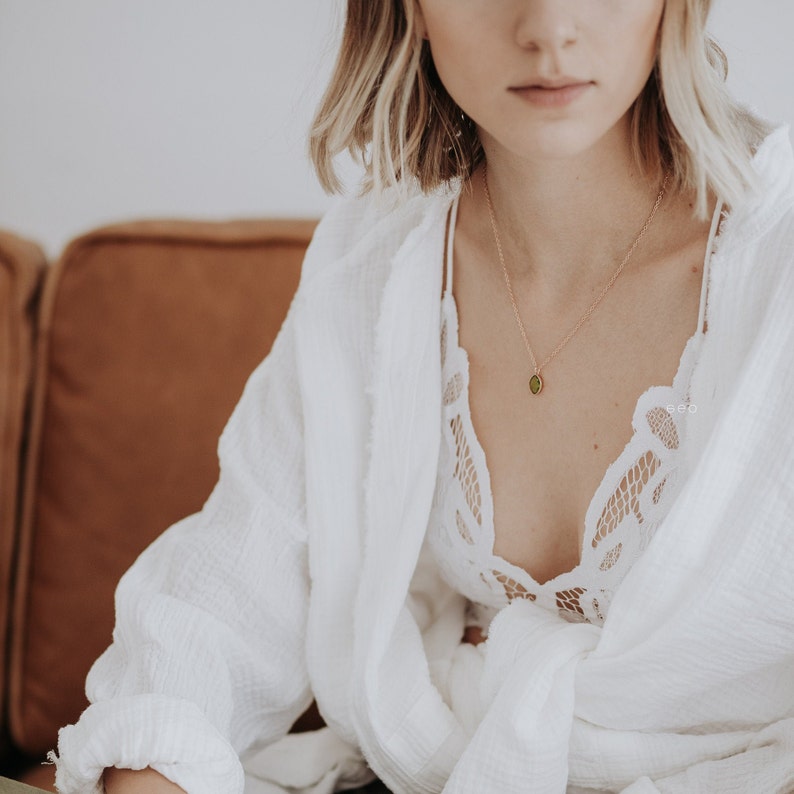 May include: A woman wearing a white shirt with a lace top underneath. She is wearing a gold necklace with a green stone pendant.