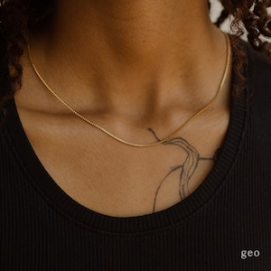 May include: A gold chain necklace on a person wearing a black shirt. The chain is a box chain style.