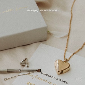 May include: Gold heart-shaped necklace with a small opening on the side. The necklace is on a white surface with a small funnel, screwdriver, and a piece of wire. The text "No longer by our side... but forever in our hearts" is written on the surface. The text "geo" is written in the bottom right corner.