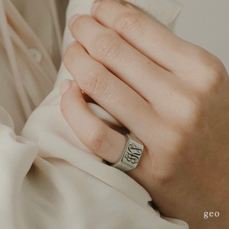 Puede incluir: Un anillo de sello de plata con una cara cuadrada que presenta un monograma. El anillo se lleva en el dedo &iacute;ndice de una mano.