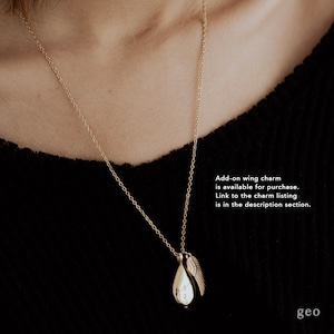 May include: A gold-coloured necklace featuring a teardrop pendant and a small wing charm. The pendant has an engraved letter. The necklace is shown against a black background. Text on the image says, "Add-on wing charm is available for purchase."