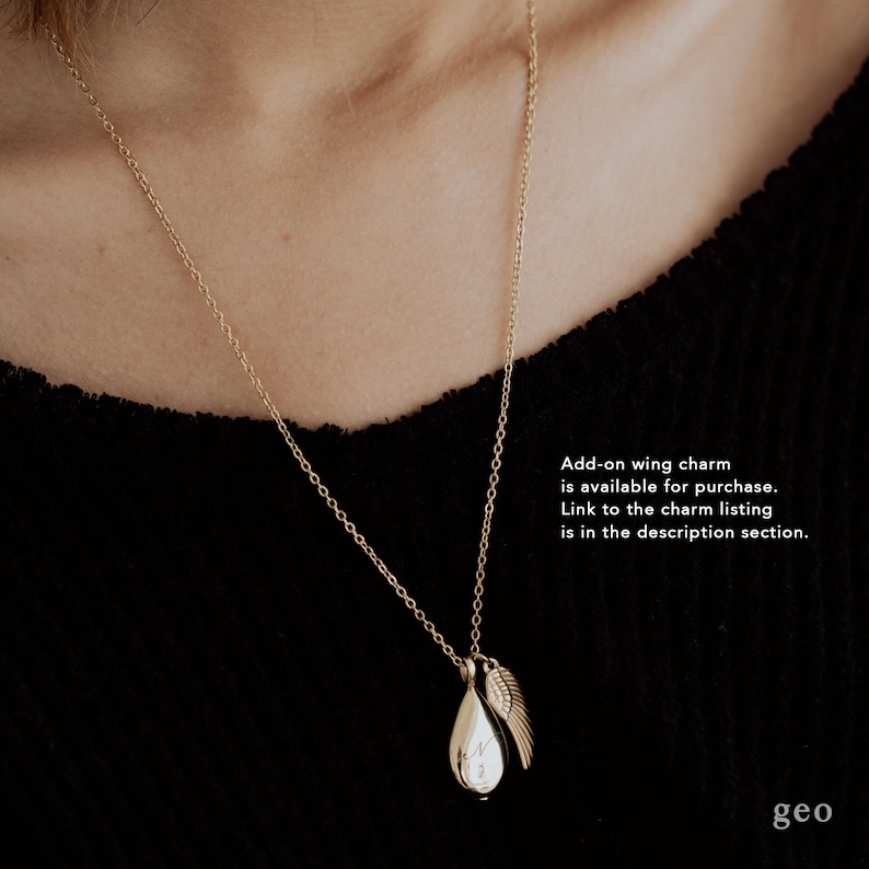 May include: A gold-coloured necklace featuring a teardrop pendant and a small wing charm. The pendant has an engraved letter. The necklace is shown against a black background. Text on the image says, "Add-on wing charm is available for purchase."