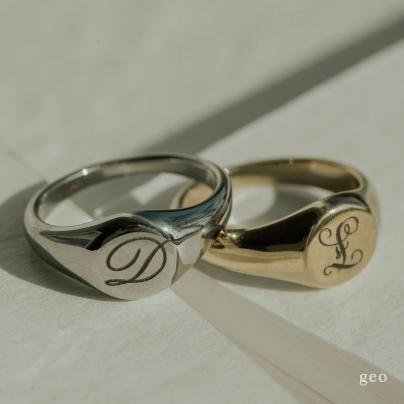 Op de afbeelding: Twee zegelringen, een zilveren en een gouden, met gegraveerde initialen. De zilveren ring heeft een 'D' gegraveerd, en de gouden ring heeft een 'L' gegraveerd.