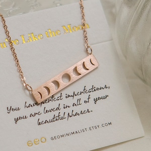 May include: A rose gold necklace with a bar pendant featuring a crescent moon design. The pendant has five cutouts representing different phases of the moon. The necklace comes with a card that reads "You have perfect imperfections, you are loved in all of your beautiful phases." and "GEOMINIMALIST.ETSY.COM".