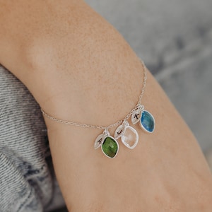 Può includere: Un braccialetto a catena d'argento con tre ciondoli. Ogni ciondolo presenta una pietra preziosa di colore diverso e una lettera incisa su una foglia. Le pietre preziose sono verde, trasparente e blu.