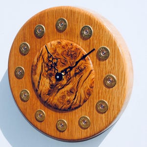 May include: A round wooden clock with a wood grain pattern. The clock face is made of a circular piece of wood with a wood grain pattern. The clock hands are black and the numbers are made of gold shotgun shells.
