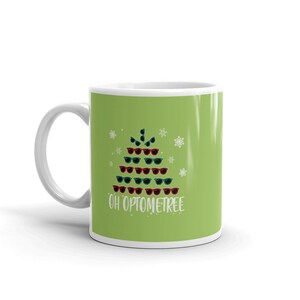 May include: A white ceramic coffee mug with a green background and a Christmas tree design made of sunglasses. The text "Oh Optometree" is printed below the tree.