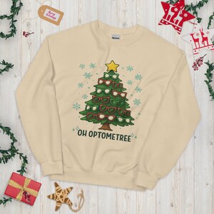 May include: A cream-colored sweatshirt features a Christmas tree design made of eyeglasses, with a yellow star on top. The phrase "OH OPTOMETREE" is printed below the tree in a playful font. Snowflake and star accents complete the festive look.