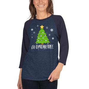 May include: A navy blue and heather gray baseball tee shirt with a green Christmas tree with eyes for ornaments. The text "OH OPTOMETREE" is printed below the tree.