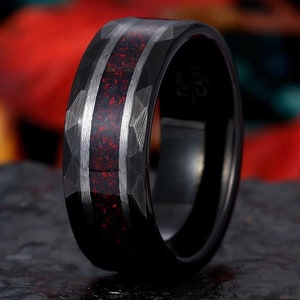 May include: A black tungsten carbide ring with a red and silver inlay. The ring has a textured, diamond-shaped pattern.