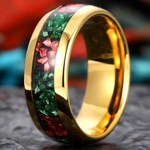 May include: A gold-coloured ring featuring a band of green and red inlaid stones. The ring has a polished finish, set against a blurred background of red and blue.