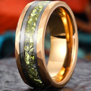 May include: A gold-colored ring with a hammered texture. The ring features a central inlay of green gemstones and a black band. The ring is displayed on a dark, textured surface, showcasing its intricate design and craftsmanship.