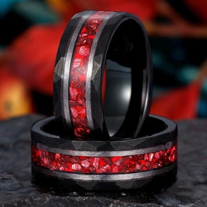 May include: Two black tungsten carbide rings with a red inlay of crushed gemstones. The rings have a hammered finish.