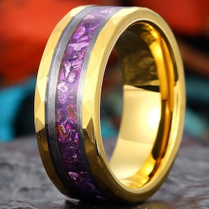 May include: Gold-colored ring with a hammered texture. The ring features a central band of crushed purple stones, bordered by a silver-colored band. The ring is set against a dark, textured surface.