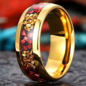 Op de afbeelding: Een goudkleurige ring met een band van rode en goudkleurige stenen. De ring heeft een gepolijste afwerking en staat tegen een vage achtergrond. De ring is een sieraad.