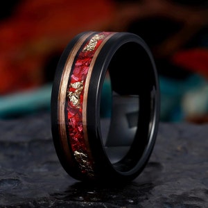 May include: Black ring with a red and gold inlay. The ring has a black band with a wood-tone and gold inlay. The center of the ring is filled with red and gold flecks. The ring is set against a dark background.